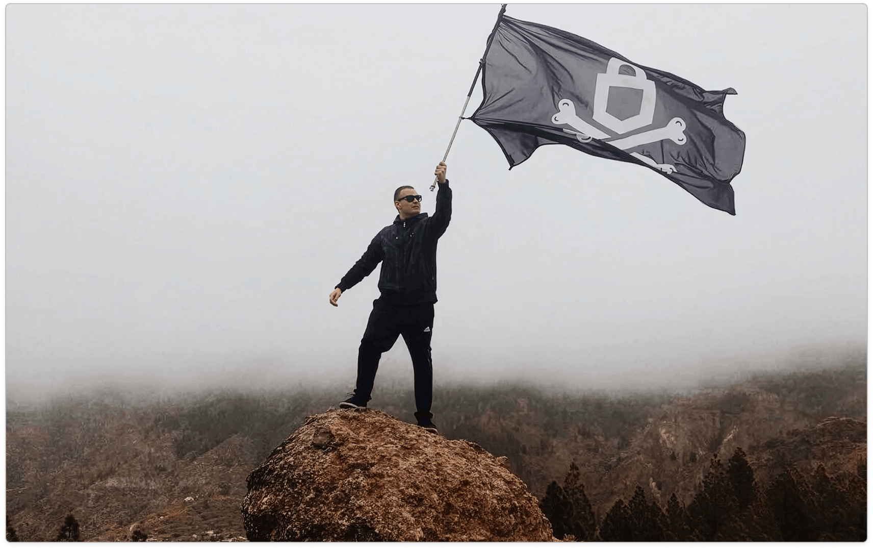 Person in black standing on a rocky peak and holding a large dark emblem flag over a hazy forested landscape.