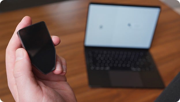 Hand holding a black screen device in front of an open laptop on a wooden desk.