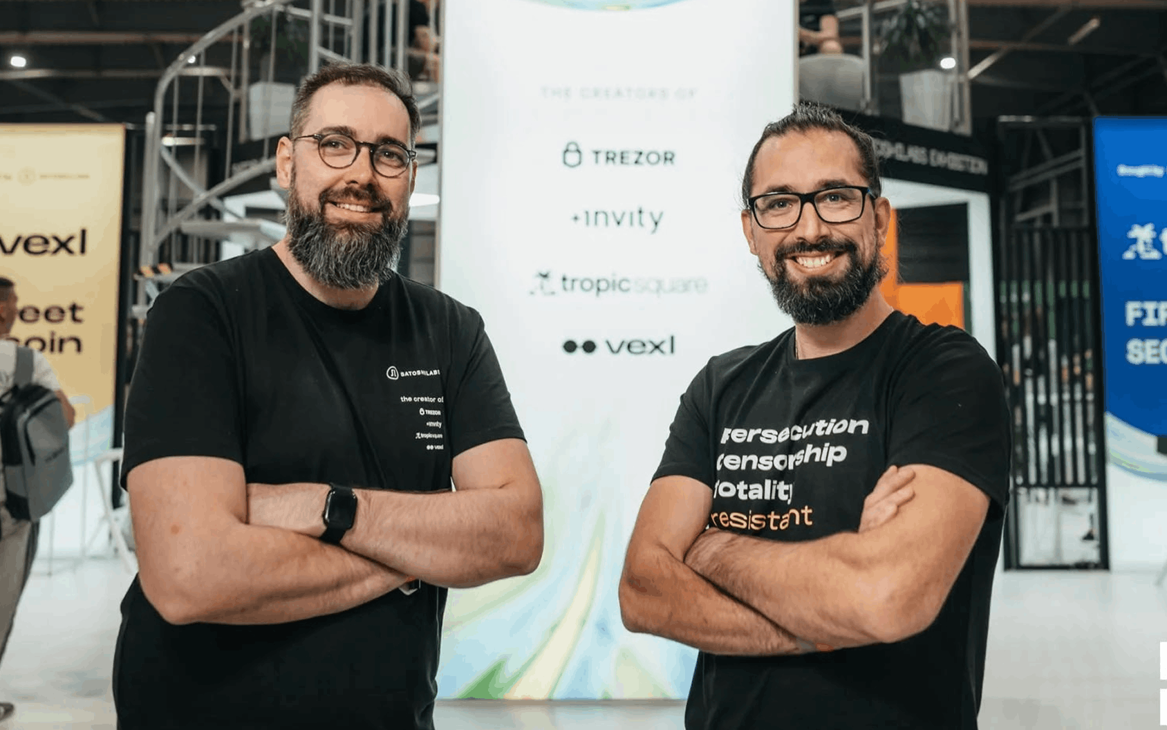 Two bearded men in black T-shirts pose with crossed arms before a sponsor backdrop at an indoor event.